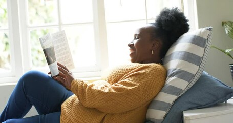 Happy african american senior woman lying, reading book and smiling in sunny room, slow motion