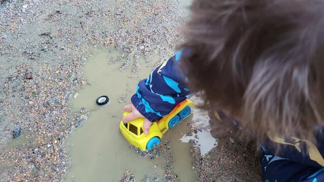 The baby is having a blast playing with toy cars in the pool of rainwater, enjoying the childhood joy of getting dirty. Kid boy aged two years (two-year-old)