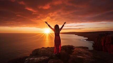 An image of a woman stands on the edge of a cliff with her arms outstretched against the backdrop of a sunset.