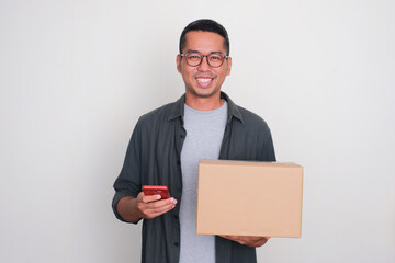 Adult Asian man smiling while holding a package box and mobile phone