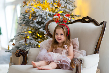 Smiling emotional little girl in an elegant dress sitting near the Christmas tree, celebrating the New Year at home. New Year's interior.