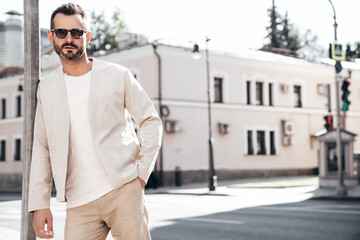 Portrait of handsome confident stylish hipster lambersexual model. Sexy modern man dressed in elegant suit. Fashion male posing in the street background in Europe city at sunset. In sunglasses - obrazy, fototapety, plakaty