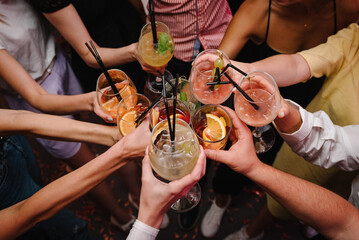Cheers with glasses at pub. Toasting, summer colorful cocktails. Happy young people cheering drinks at bar restaurant. Party time concept. Group of friends partying in nightclub. Top view of hands up - obrazy, fototapety, plakaty