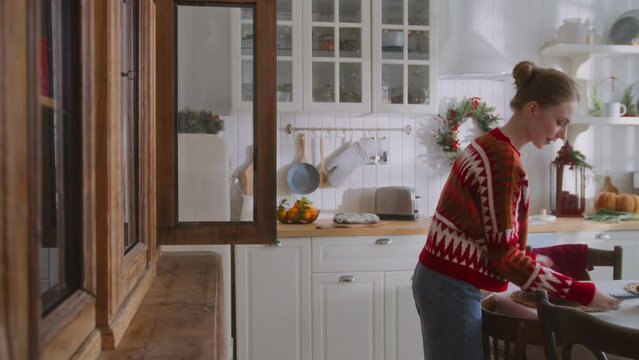 young woman takes plates out of buffet to serve table for christmas dinner at daylight in kitchen. pick up phone call, gets great news, becomes happy and surprised, smiles and laughs unbelieving