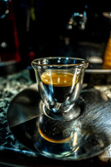 Macro view of a modern designed glass cup with freshly brewed black coffee with golden foam with small bubbles perched on a metal coffee plate on top of a barista automatic espresso machine.