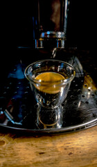 Modern design glass cup of glass with freshly brewed black coffee with golden foam with small bubbles on top of an automatic barista espresso machine on a rustic wooden table.