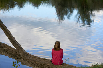 Rear view of a woman in a pink sweater sitting on a fallen tree in front of the river in morning contemplating the landscape relaxedly. Concept: connection with nature and outdoor lifestyle