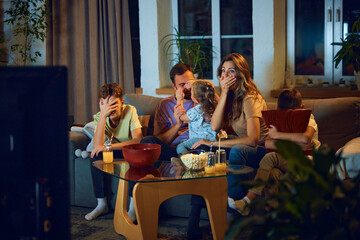 Family watching tv at home in the evening. Parents with children sitting on couch, watching horror movie, covering eyes in fear. Concept of family, leisure time, relaxation, childhood and parenthood - obrazy, fototapety, plakaty