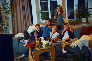 Happy loving family, parents and children sitting on couch in the evening at home and watching tv, eating snacks. Relaxation. Concept of family, leisure time, relaxation, childhood and parenthood