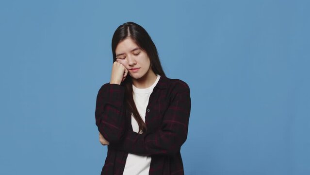 Young attractive woman falls asleep standing, resting her head on her hand and then suddenly wakes up with a surprised face on a blue background in the studio