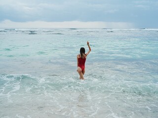 Woman standing with her back to the camera tourist in a red bathing suit sitting on the sand on the beach in the ocean in the waves, travel and beach vacation