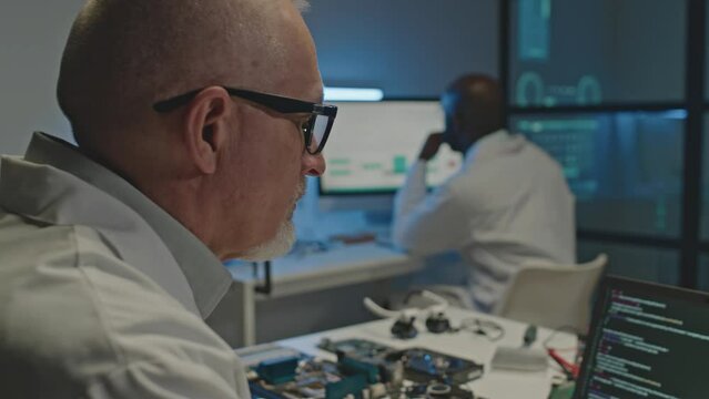 Waist up of mature Caucasian male electronics engineer program coding on laptop while sitting at desk with circuit board parts on it in dark lab, working there with African American male colleague
