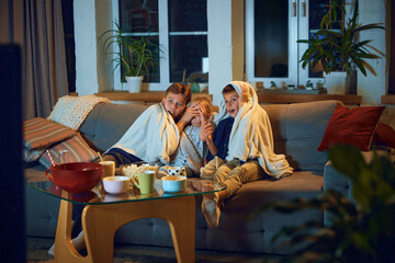 Emotional children, boys and little girl sitting on couch at home and watching tv, horror movie. Hiding under blanket. Concept of family, leisure time, relaxation, childhood and parenthood