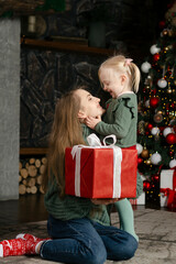 Obraz na płótnie Canvas Happy young mother and little daughter embrace near Christmas tree with large Xmas present. Vertical frame.