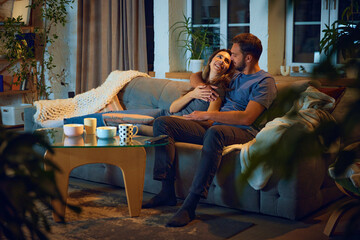 Loving young couple, man and woman sitting on sofa in evening at home,, hugging, cuddling and...