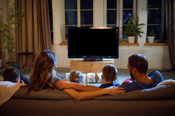 Back view of parents and children spending evening together, sitting on couch in living room and...