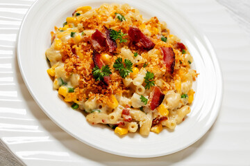 close-up of portion of macaroni and cheese with corn, bacon topped with panko breadcrumbs on white plate on white wooden table, dutch angle view
