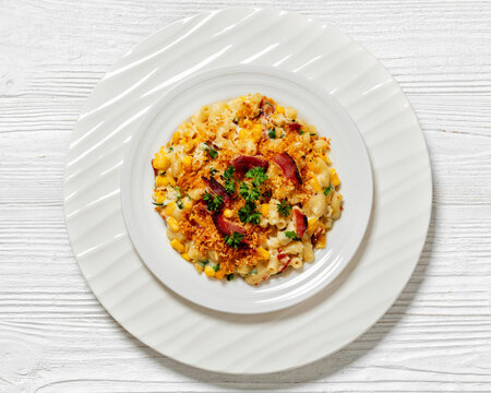 portion of mac and cheese with corn, bacon topped with panko breadcrumbs on white plate on white wooden table, horizontal view from above, flat lay, close-up