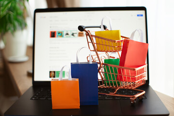 E-commerce and online shopping concept. Shopping cart, laptop, colourful bags near the window. Blurred background.