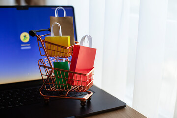 E-commerce and online shopping concept. Shopping cart, laptop, colourful bags near the window. Blurred background.