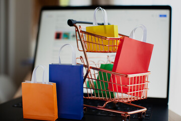E-commerce and online shopping concept. Shopping cart, laptop, colourful bags near the window. Blurred background.