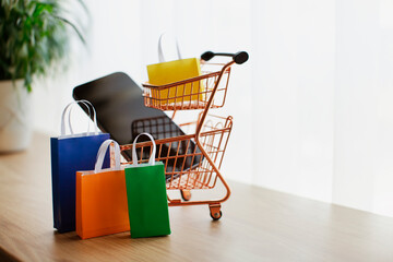 E-commerce and online shopping concept. Shopping cart, mobile phone, colourful bags near the window. Blurred background.