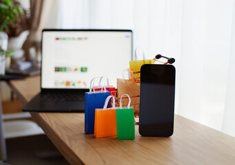 E-commerce and online shopping concept. Laptop, shopping cart, mobile phone, colourful bags near the window. Blurred background.