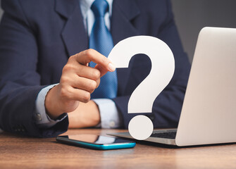 Businessman holds a white question mark symbol sitting at the table.