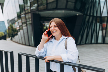 Young happy redhead businesswoman making phone call over mobile phone in the city. Middle age woman talking on smartphone, walking city urban street at business office center. Manager work outside