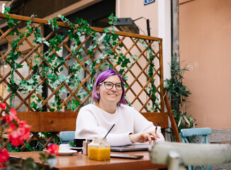 Stylish woman with purple hair sitting in the cafe's garden and having a drink while working