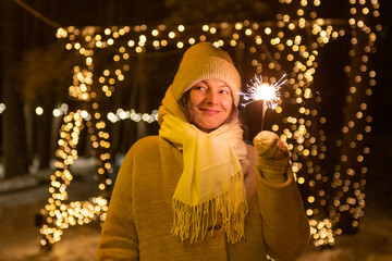 Young woman holding burning sparklers against unfocused festive lights (selective focus)