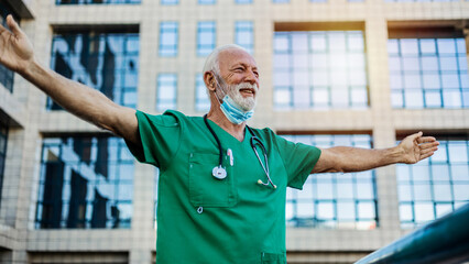 A mature doctor is standing in front of the hospital with open arms and feeling relieved after a...