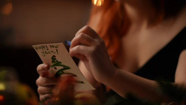 Close-up. Red-haired woman signing or drawing on the back of a New Year's card with a ballpoint pen