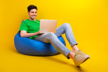 Full size photo of clever positive guy wear t-shirt sit on bean bag look at laptop chatting at work...