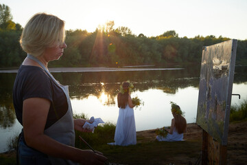 Adult female artist painting picture near water of river or lake in nature and girls in white...