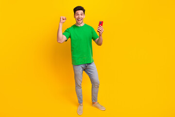 Full size photo of impressed guy wear stylish t-shirt denim pants hold smartphone win gambling isolated on yellow color background