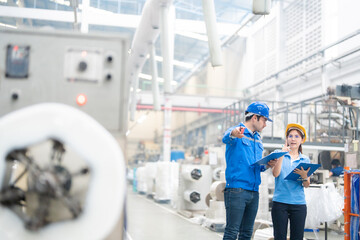 side view photo In factories that produce plastic and steel There's an American male engineer and an Asian female manager pointing at working machinery, wearing uniforms, hard hats, and lisnots.