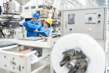 American male engineer and Asian female manager Inspecting work systems in plastic and steel industries Wearing a uniform and a hard hat, holding a list of notes, machinery is working around.