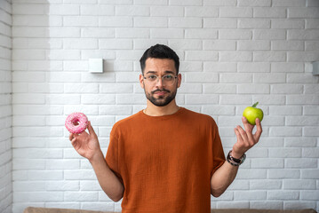 Young student man doubts what to choose healthy food or sweets junk unhealthy food holding green apple and donuts in hands standing at home in her living room. Hard choice concept eating disorder - obrazy, fototapety, plakaty