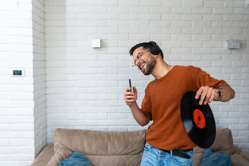 Young happy man singing and dancing at home listening music on wireless headset singing on his phone holding LP record in his hand. Hipster music lover digitized old record audio and enjoying - obrazy, fototapety, plakaty