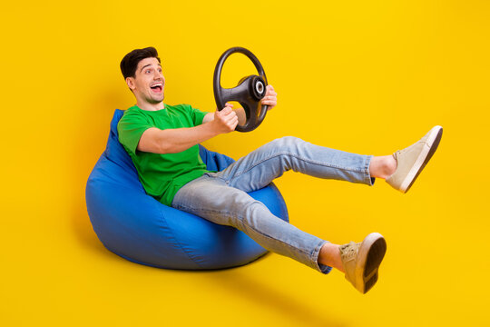 Full size photo of ecstatic guy wear t-shirt sit on bean bag look empty space hold steering wheel isolated on yellow color background