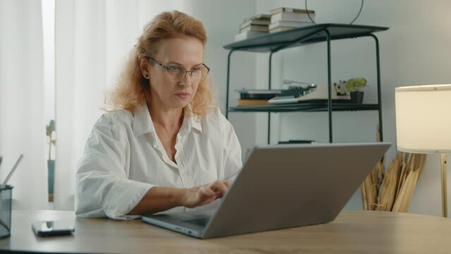 Attractive mature blonde in white T-shirt with rolled sleeves working on silver laptop, deleting the text sitting at table at home in a cozy light room. High quality 4k footage