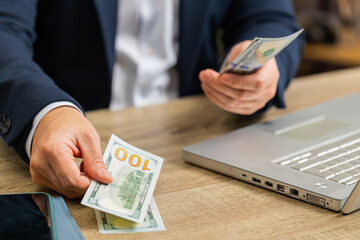 Close-up of hands of rich businessman counting money hundred dollar cash income use laptop at home office workplace table. Professional freelancer man rejoicing win, success contract, earning profit