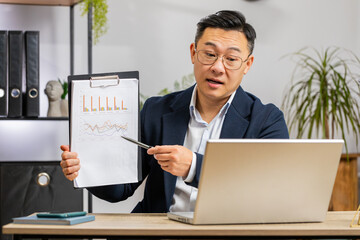 Asian businessman freelancer making online presentation video laptop call education lessons for remote study student classes at office workplace. Male Chinese in suit demonstrates graphic analytics