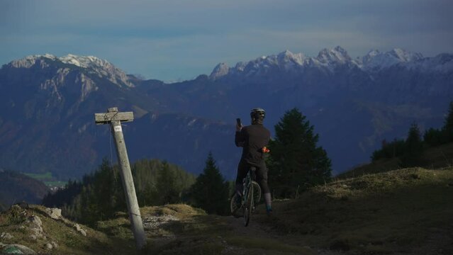 Male cyclist on gravel bike enjoying view and making phono on phone on top of mountain in alps with beautiful view of snowy peaks of mountain range in germany. Sports motivation and inspiration. 