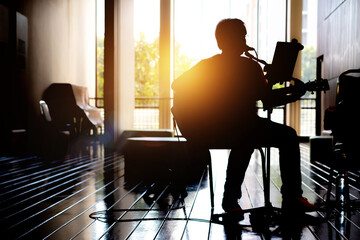 Silhouette of a man sitting on a chair playing a electric guitar in hall hospital and  background of the sunset. Silhouette romance concept, music therapy - Powered by Adobe