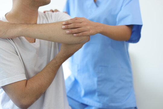Doctor doing physiotherapy and diagnosing male patient arm and shoulder pain in hospital examination room.