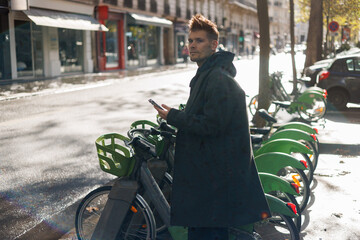 Young man unlocks bicycle with his mobile phone. Electric bicycle new way city mobility. Green...
