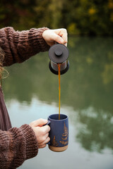 Man with long hair pouring coffee in a mug outside in nature by the river