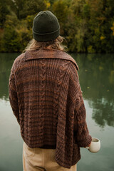 man with long hair in a whool coat and beanie holding a cup of coffee and relaxing looking forward at the river.